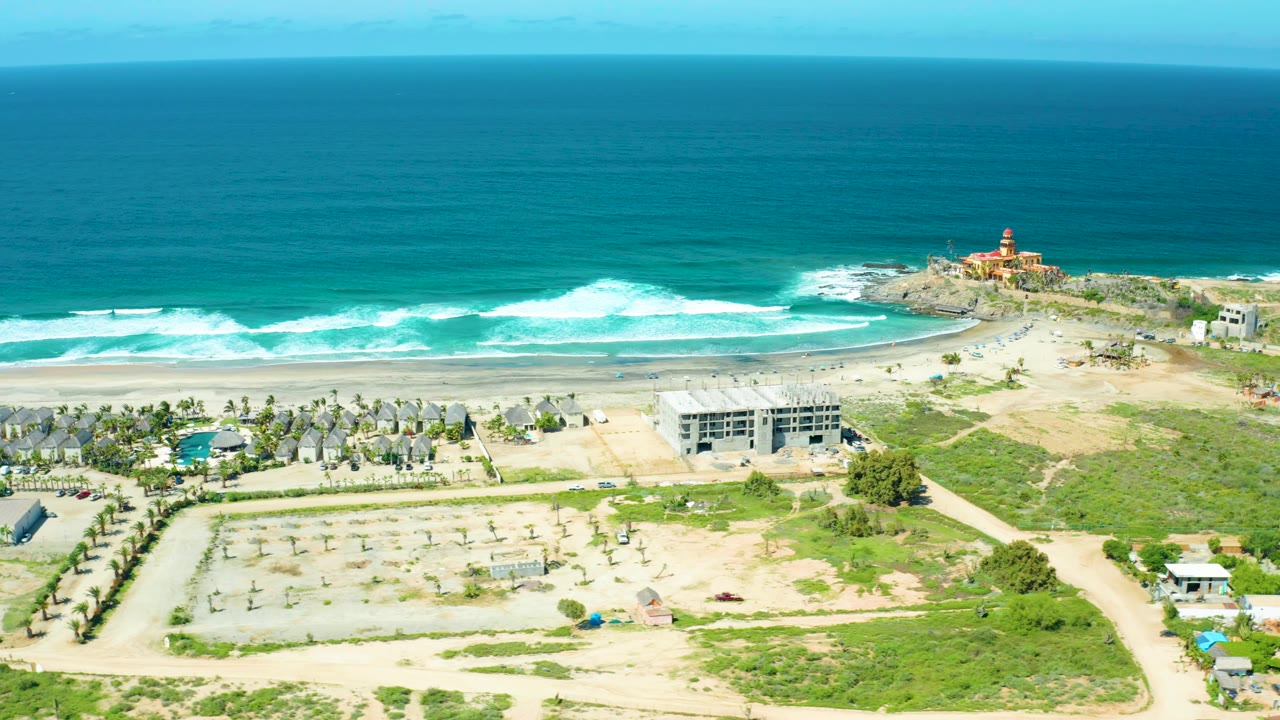 Cerritos Beach aerial frame with a fresh line of white surf, sandy beach, clustered temporary structures, dirt lots, a few low buildings, a rocky point, and no visible people.