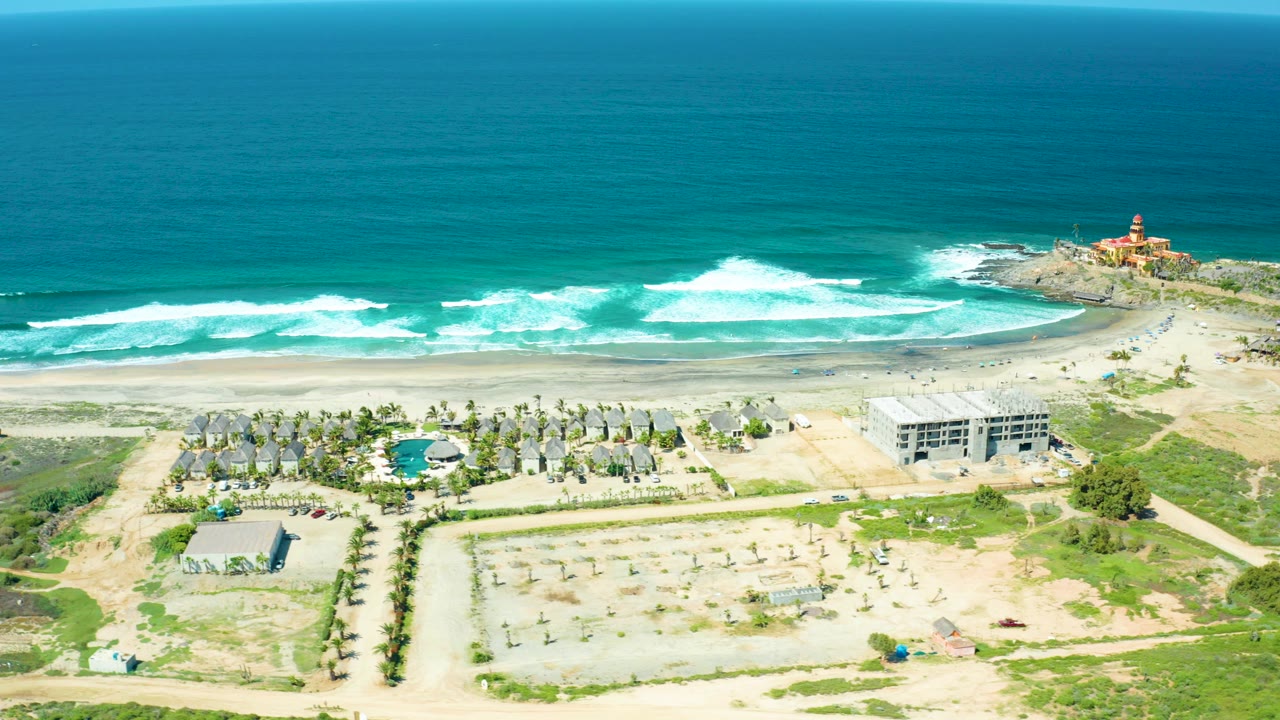 Aerial view of ocean waves, beach resort buildings, dirt lots, and a small lighthouse.
