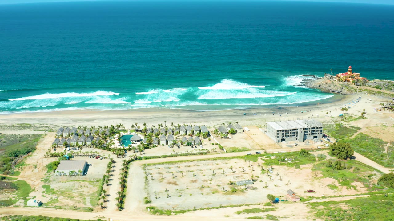 A wide aerial view at Cerritos Beach shows the resort compound and pool below the surf zone, with a pale sand beach, open ocean, dirt access roads, and lighthouse visible to the right, no visible people.