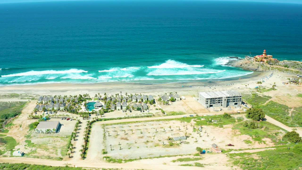 Cerritos Beach appears from above with blue-green water and white wave bands behind resort grounds, a rectangular unfinished building, pale construction lots, scrub vegetation, and the lighthouse near the right edge, no visible people.