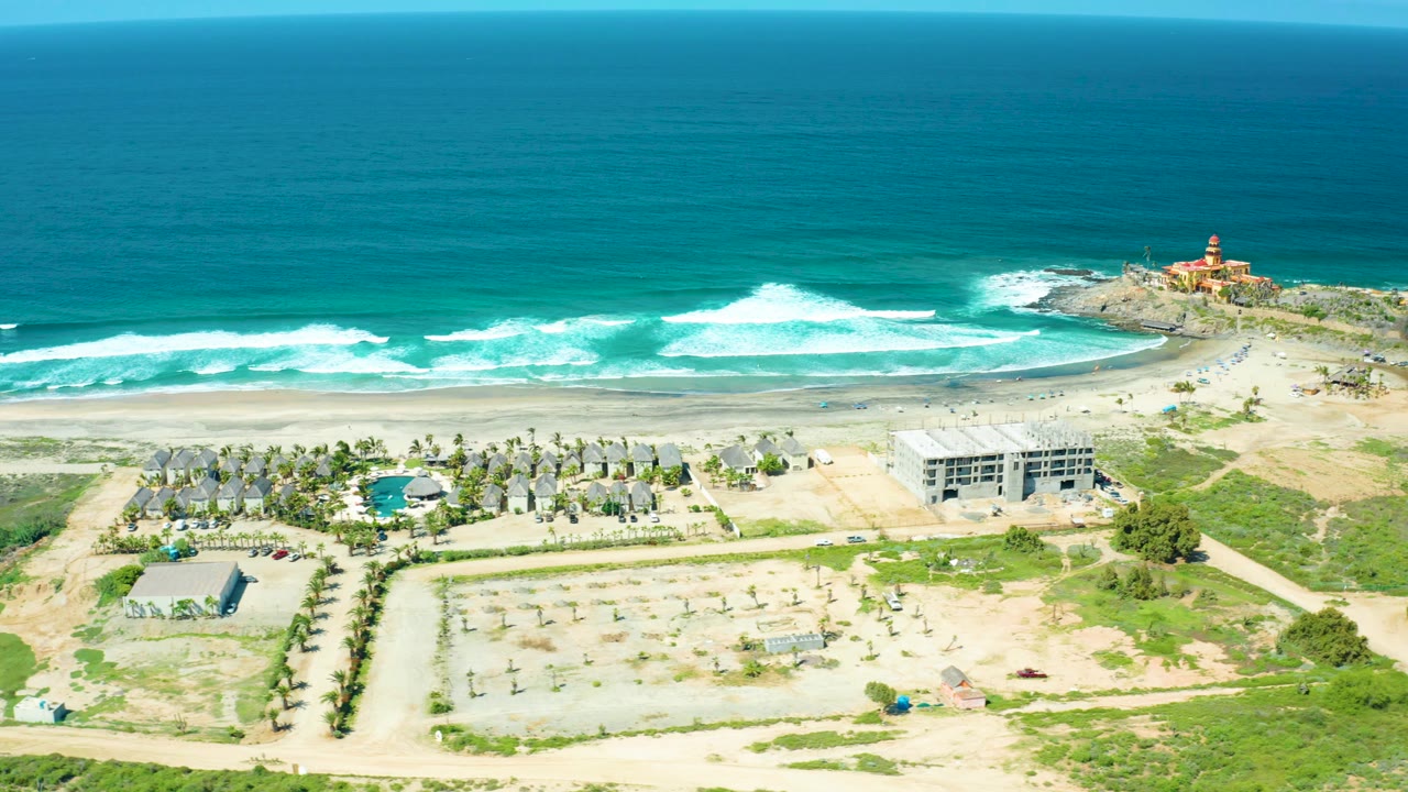 A high coastal composition over Cerritos Beach places resort buildings, a pool, dirt roads, and scrub in the lower frame, with the sand beach, bright surf, ocean horizon, and lighthouse beyond, no visible people.