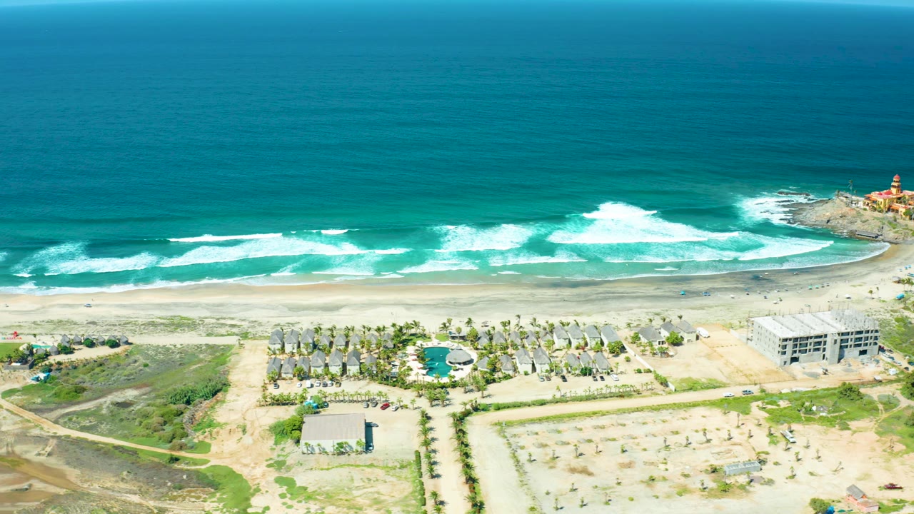 Wide drone view of waves breaking across a sandy beach with palms and low buildings inland.