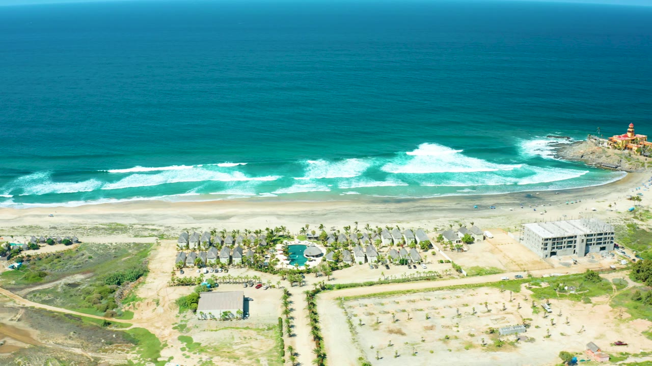 At 0:02, Cerritos Beach appears from above with foamy surf crossing the shore, a pale sand strip, palm-lined grounds, a circular blue pool-like feature, nearby buildings, and a small lighthouse at the rocky edge, no visible people.
