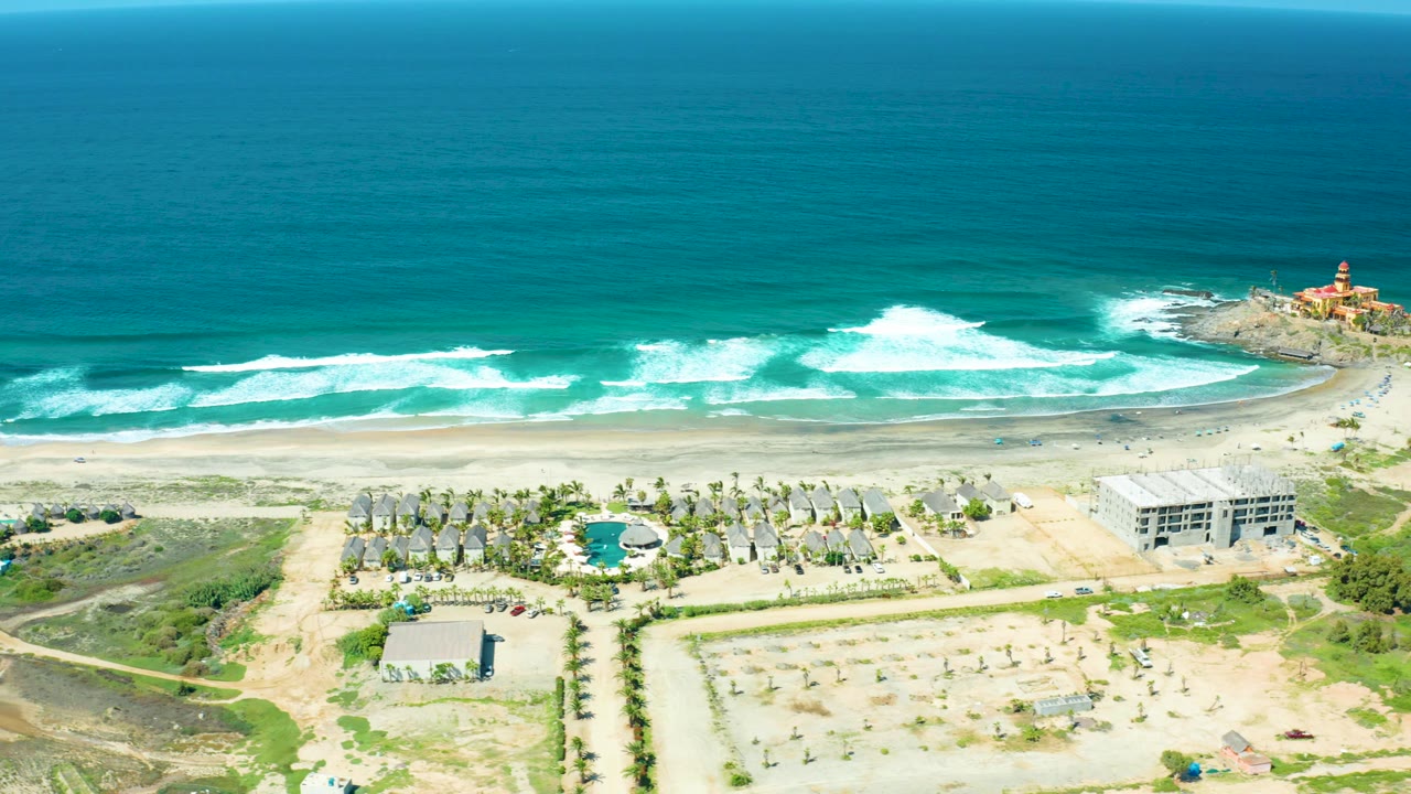 High aerial view of ocean waves, beach, palms, coastal lots, buildings, greenery, and lighthouse.