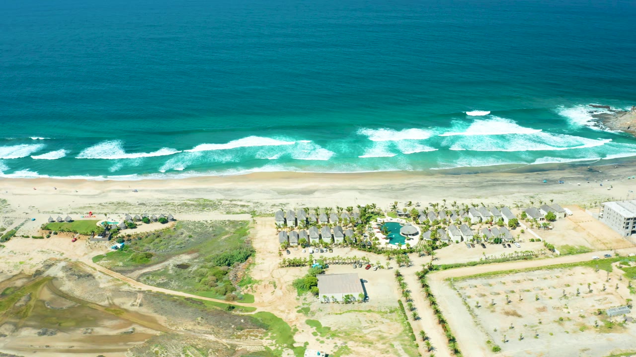 Cerritos Beach appears from above with surf bands slightly shifted along the shoreline, a pool and palm rows in the foreground, open construction ground, and no visible people.