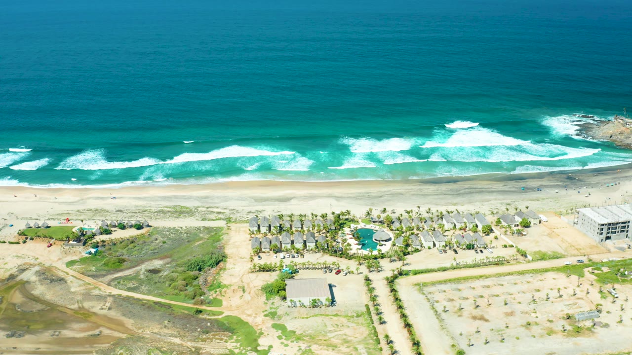 Aerial beach scene with large ocean area, breaking waves, palms, buildings, and dirt lots.