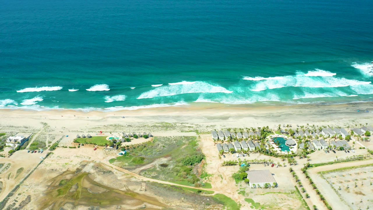 High aerial view over Cerritos Beach, Baja California Sur, with turquoise ocean, white surf, sandy shore, a pool area, roads, palms, and no visible people.