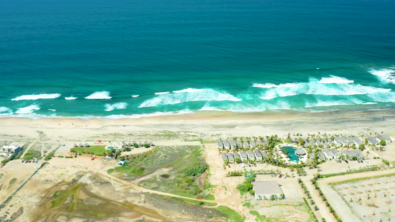 Drone-style overhead view at Cerritos Beach showing turquoise water, white foam lines, pale sand, a palm-lined pool area, low buildings, roads, and no visible people.