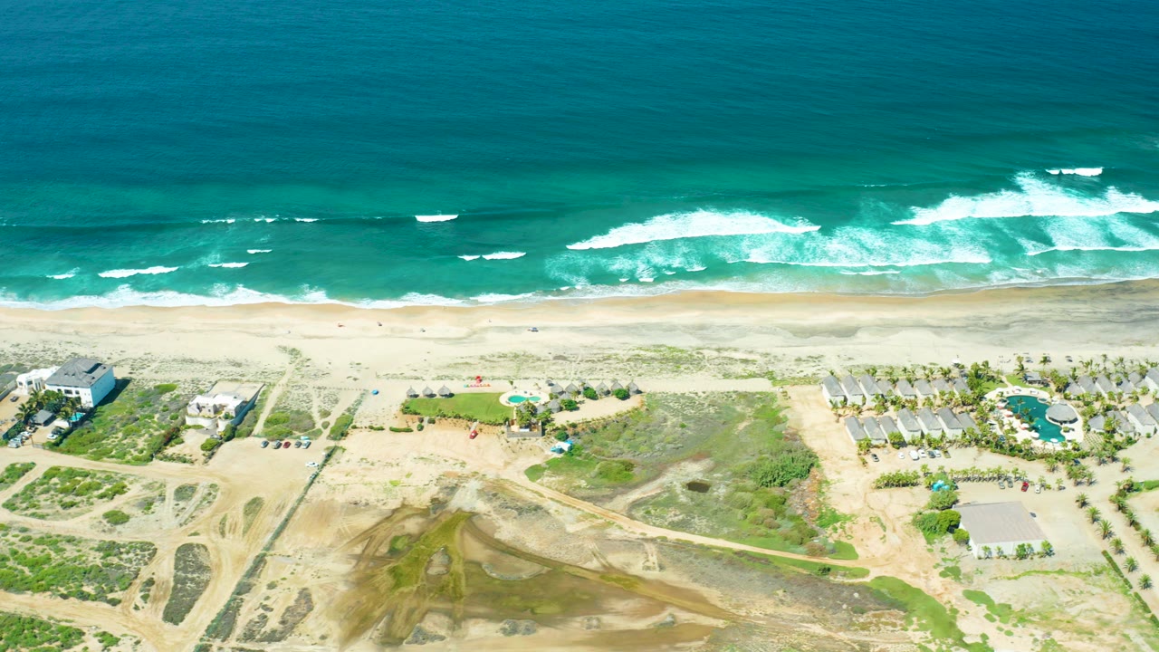 Aerial frame over Cerritos Beach, Baja California Sur, with turquoise ocean at the top, white surf bands on the sand, scattered tiny beachgoers, dunes, houses, and a round pool inland.