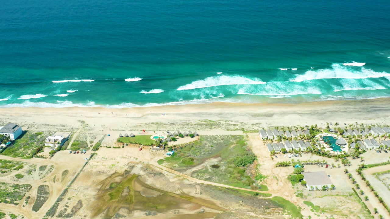 Overhead beach view with rolling surf, sand, dunes, dirt roads, and homes.