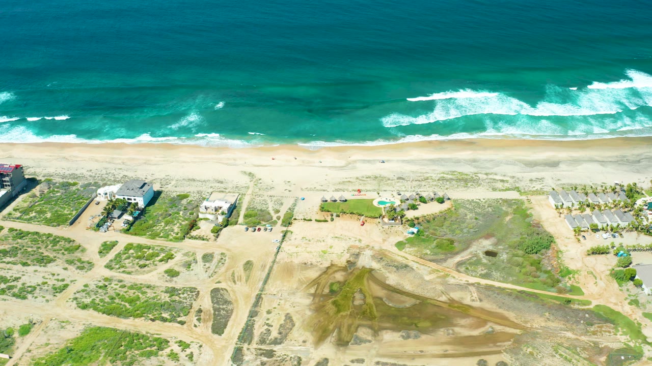 Drone view of ocean waves breaking beside a sandy beach and dune paths.