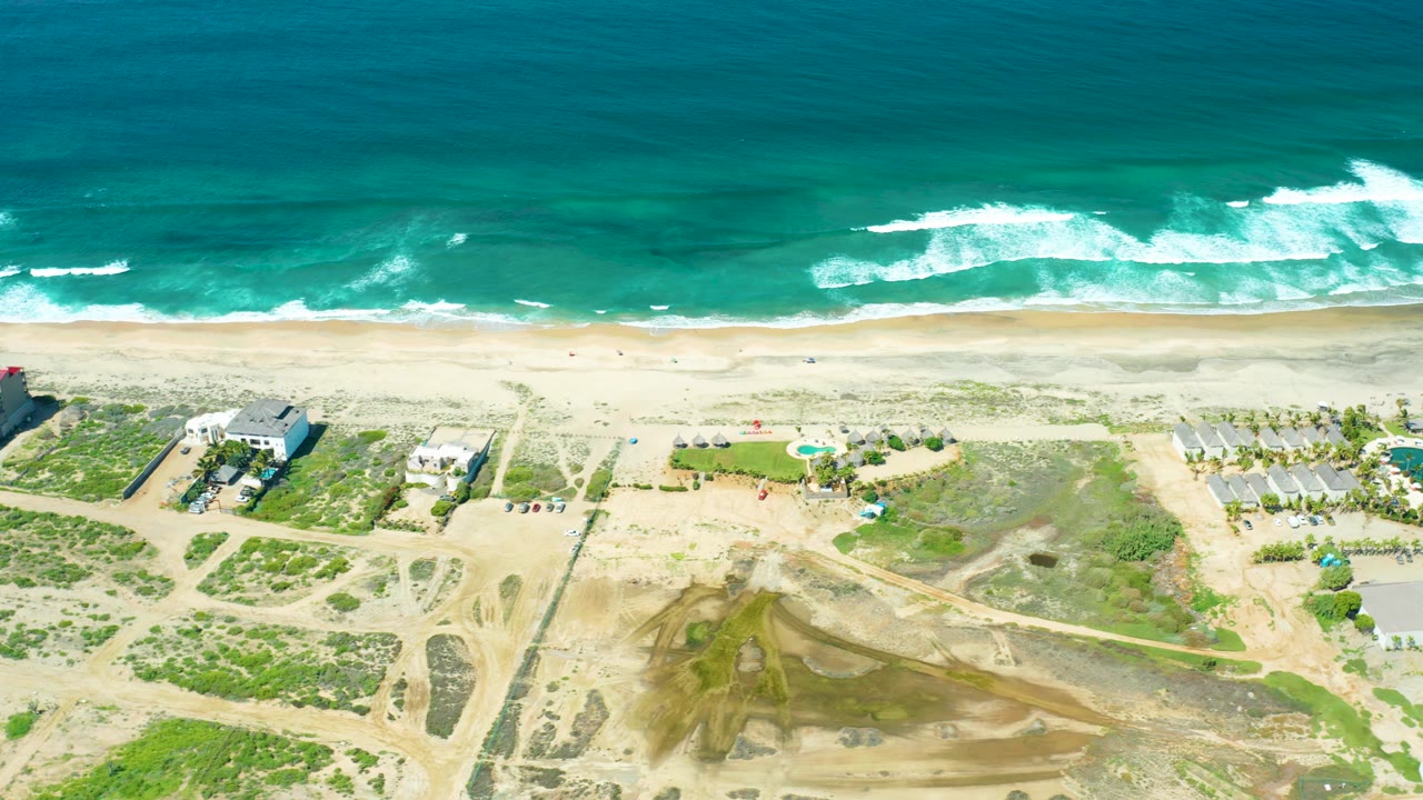 High drone view over Cerritos Beach with whitecaps across the teal ocean, a narrow sandy shore, dune vegetation, visible pools, dirt tracks, scattered buildings, and no visible people.