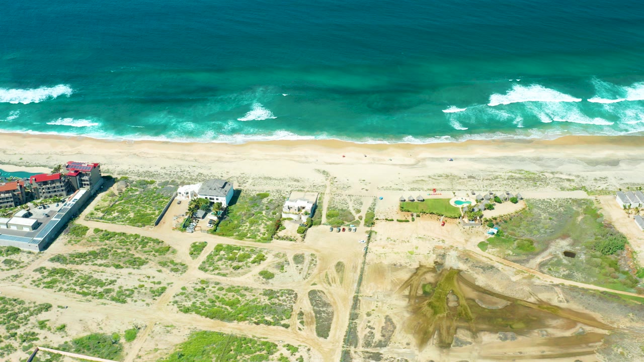 Aerial frame over Cerritos Beach, Baja California Sur showing turquoise surf, pale shoreline, sandy dunes, dirt paths, and low coastal buildings with no visible people.