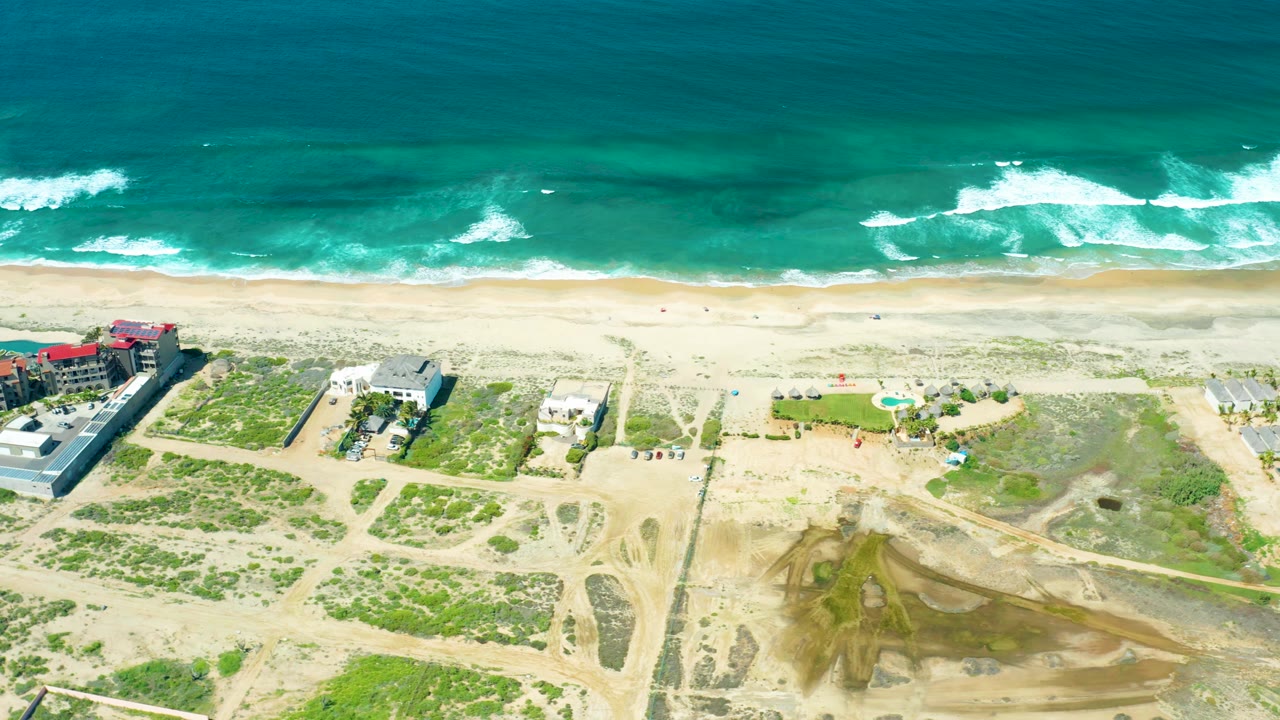 A high aerial view over Cerritos Beach shows active-looking sandy tracks, dune patches, scattered buildings, beach sand, and rolling ocean surf with no visible people.