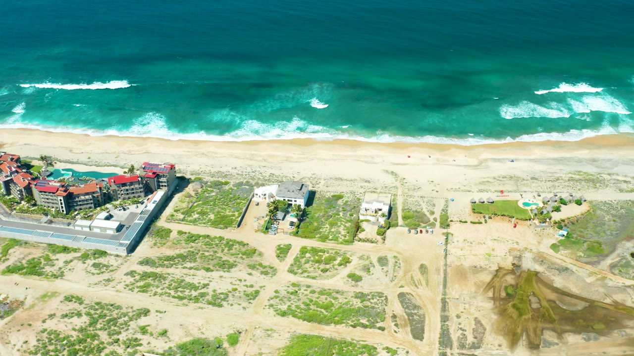 Cerritos Beach aerial frame with white wave foam shifting along the upper shoreline, pale sand in the center, inland dune tracks below, red-roofed buildings at left, and no visible people.