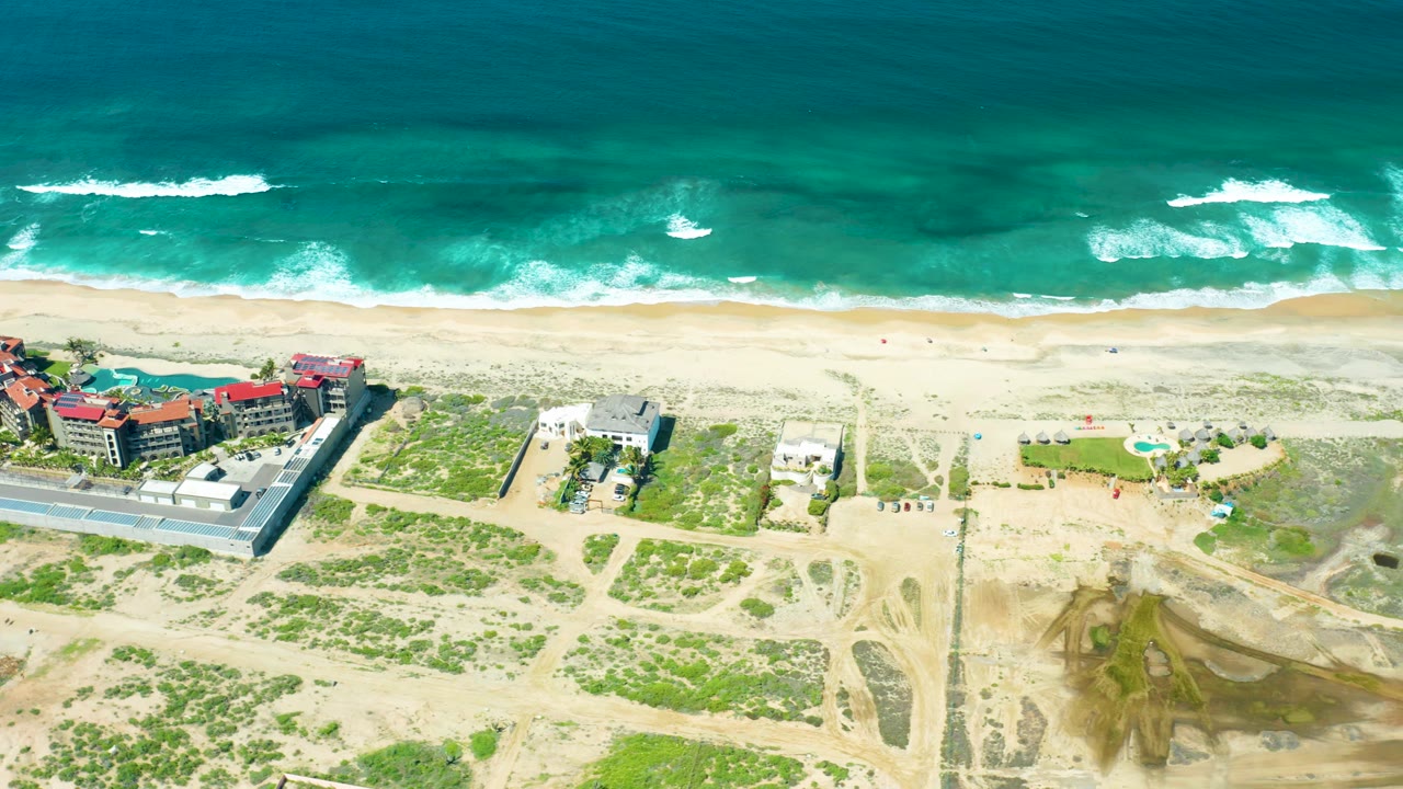 Overhead Cerritos Beach view with broader white surf bands near the top, a clean sandy strip through the middle, sparse dune vegetation below, red-roofed structures left of center, and no visible people.