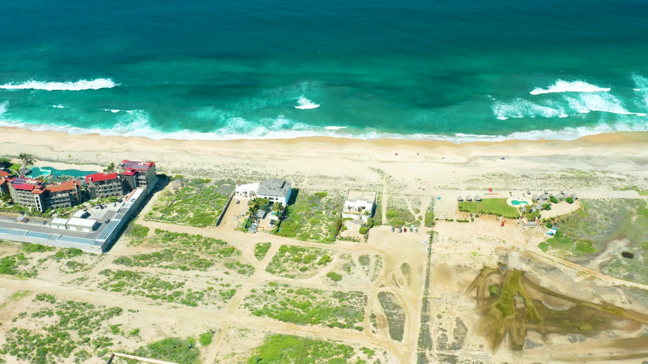 Waves break near a sandy beach with dunes, paths, and beachfront buildings below.