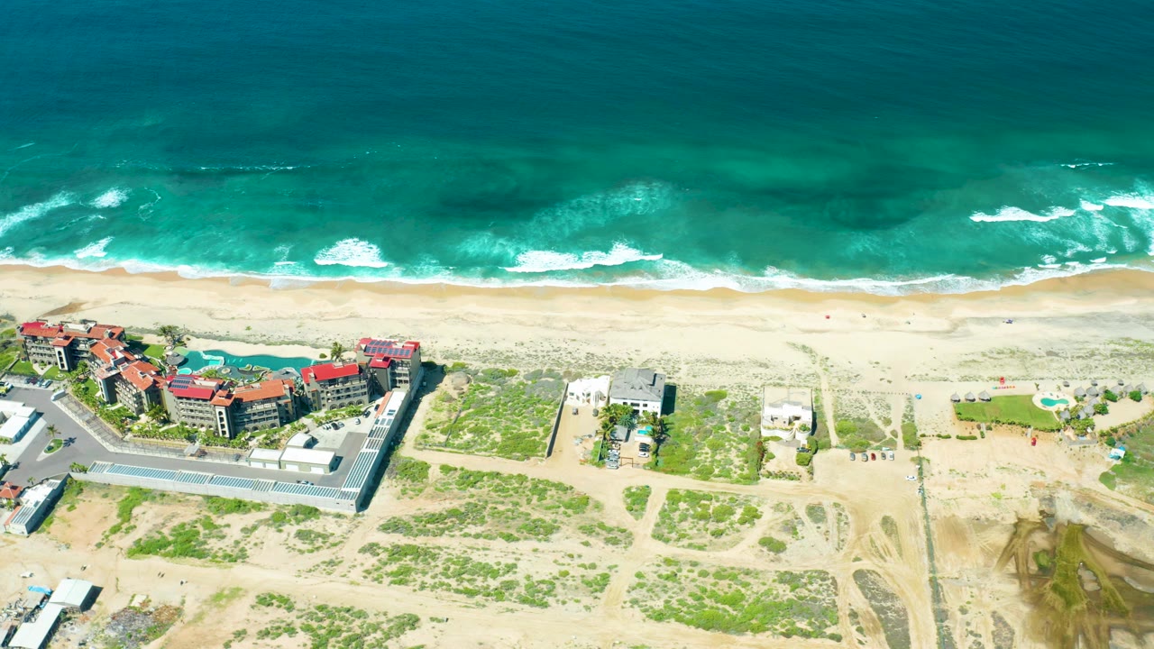 Overhead beach view with breaking waves, sandy shore, buildings, roads, and scrub dunes.
