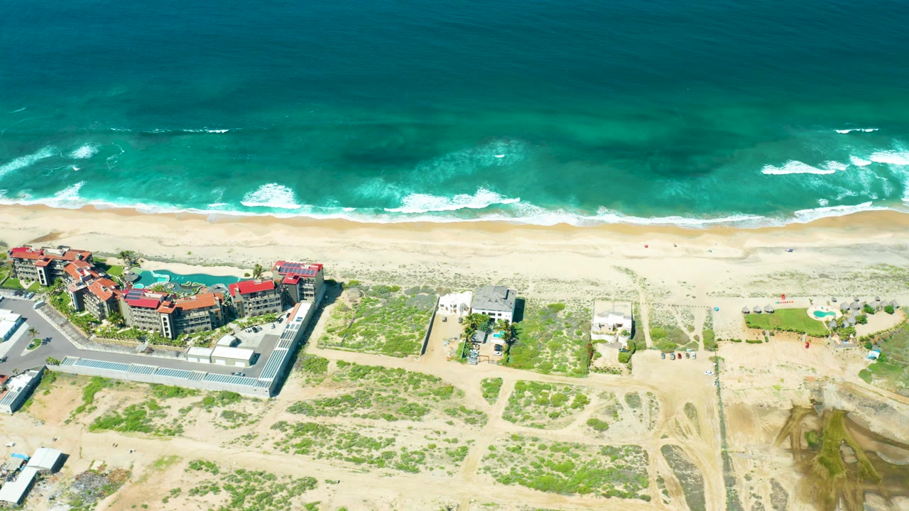Cerritos Beach aerial frame with whitecaps further along the shoreline, tiny beachgoers on the sand, clustered red-roofed buildings, a paved road, dunes, and dirt access tracks.