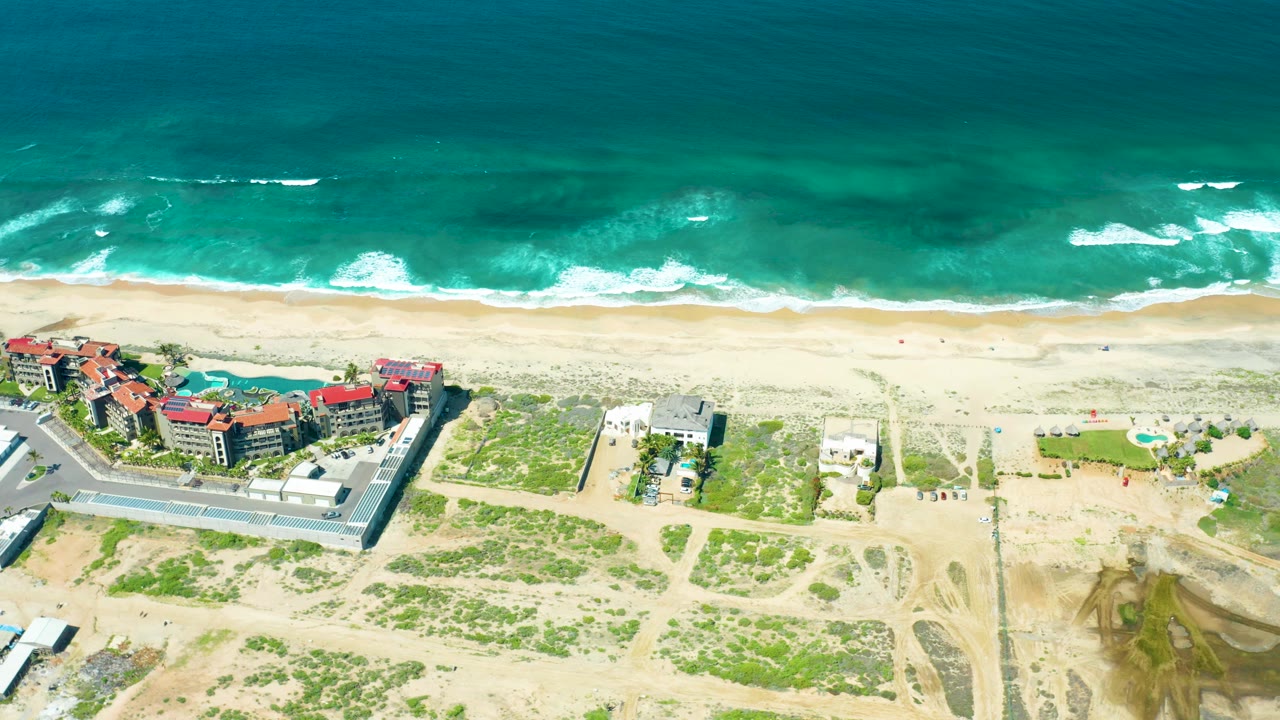 High aerial beach frame with ocean, surf, sand, buildings, road, and dune vegetation.