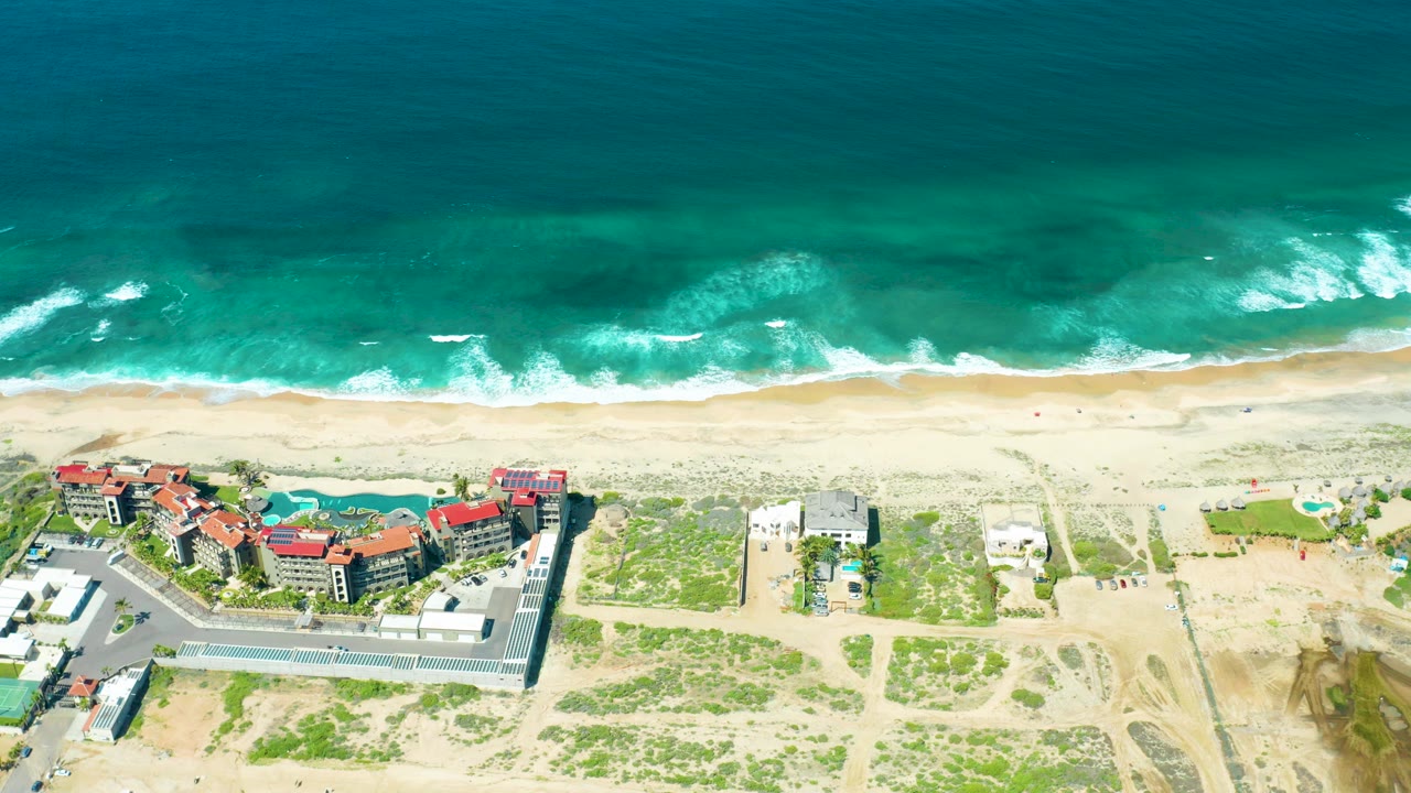 Aerial frame from Cerritos Beach, Baja California Sur, showing turquoise waves, white foam, pale sand, coastal buildings, a pool, parking areas, and no visible people.