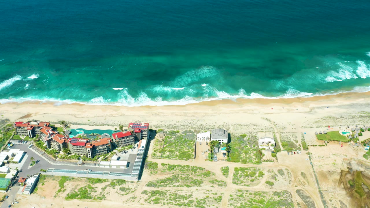 Overhead beach view with surf foam, sand, resort-style buildings, pool, and parking lot.