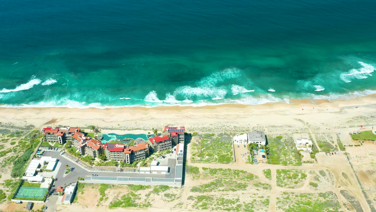 At 0:00, the Cerritos Beach aerial shows turquoise water and white surf above a pale sand beach, with red-roofed coastal buildings, a pool area, parking, dunes, and tiny people near the shoreline.
