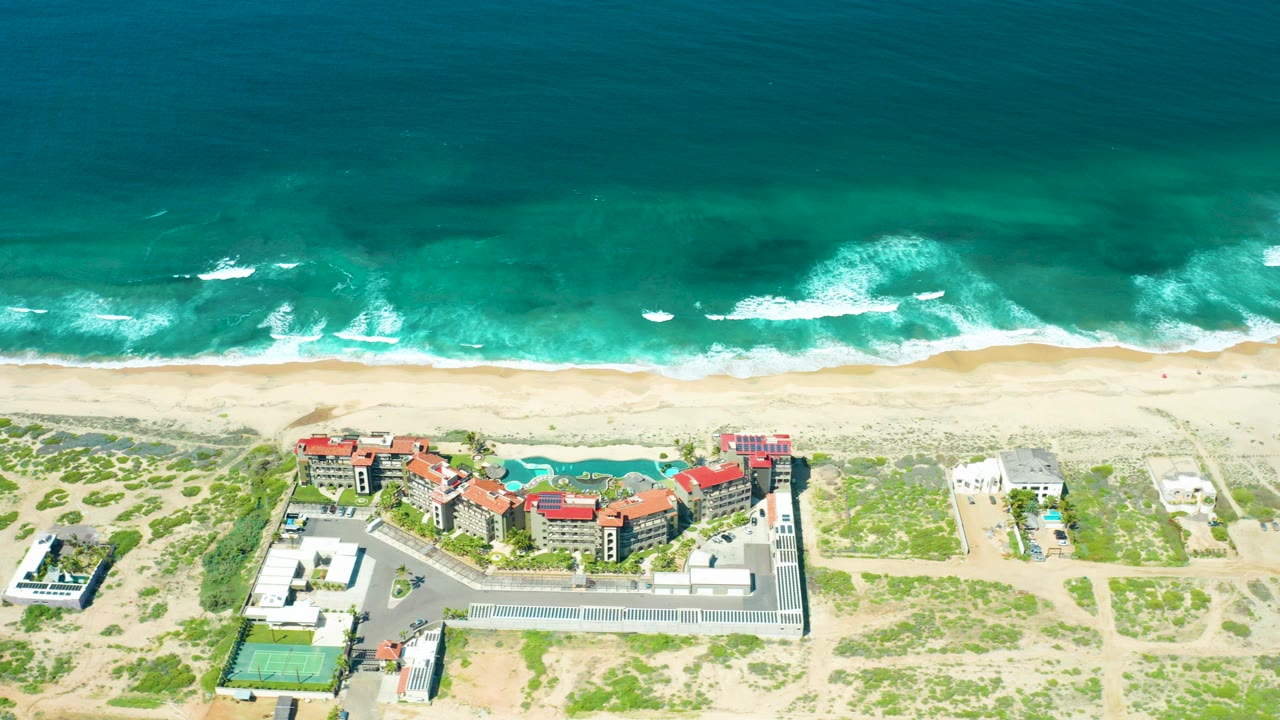 Opening aerial frame over Cerritos Beach, Baja California Sur, with turquoise waves breaking onto sand, red-roofed coastal buildings below, and no visible people.