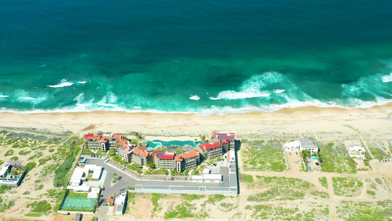 Cerritos Beach aerial frame with darker blue-green water offshore, foamy breakers along the sand, dune vegetation, red-roofed coastal buildings, and no visible people.