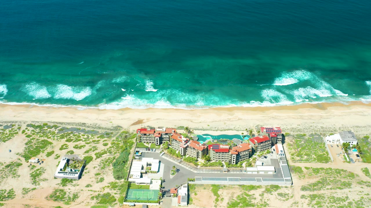 Aerial view of turquoise surf, sandy beach, dunes, and red-roofed coastal buildings.