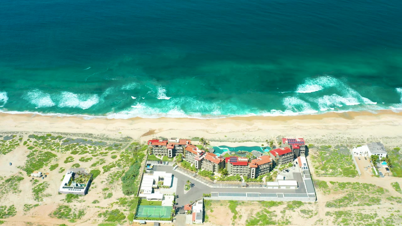 Aerial frame at Cerritos Beach showing whitecaps moving closer to the sand, a wide dune strip, red-roofed beachfront buildings, a pool, parking area, tennis court, and no visible people.