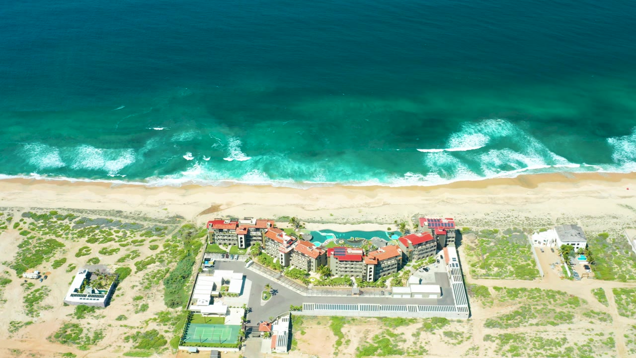Mid-clip aerial view of Cerritos Beach with teal ocean water, foamy wave breaks, a pale beach, low dune paths and greenery, red-roofed coastal buildings, a parking lot, tennis court, and no visible people.