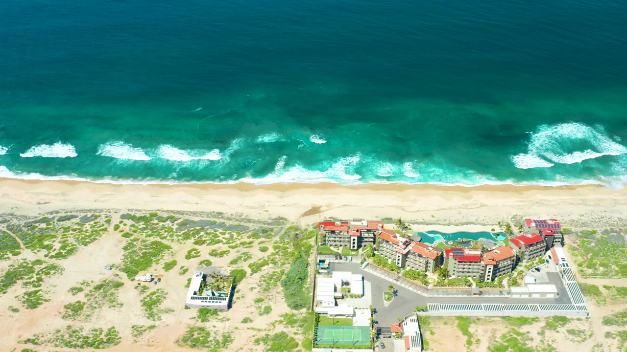 High aerial frame over Cerritos Beach, Baja California Sur, Mexico, with turquoise water, white foam bands, pale sand, dunes, red-roofed buildings, parking areas, and no visible people.