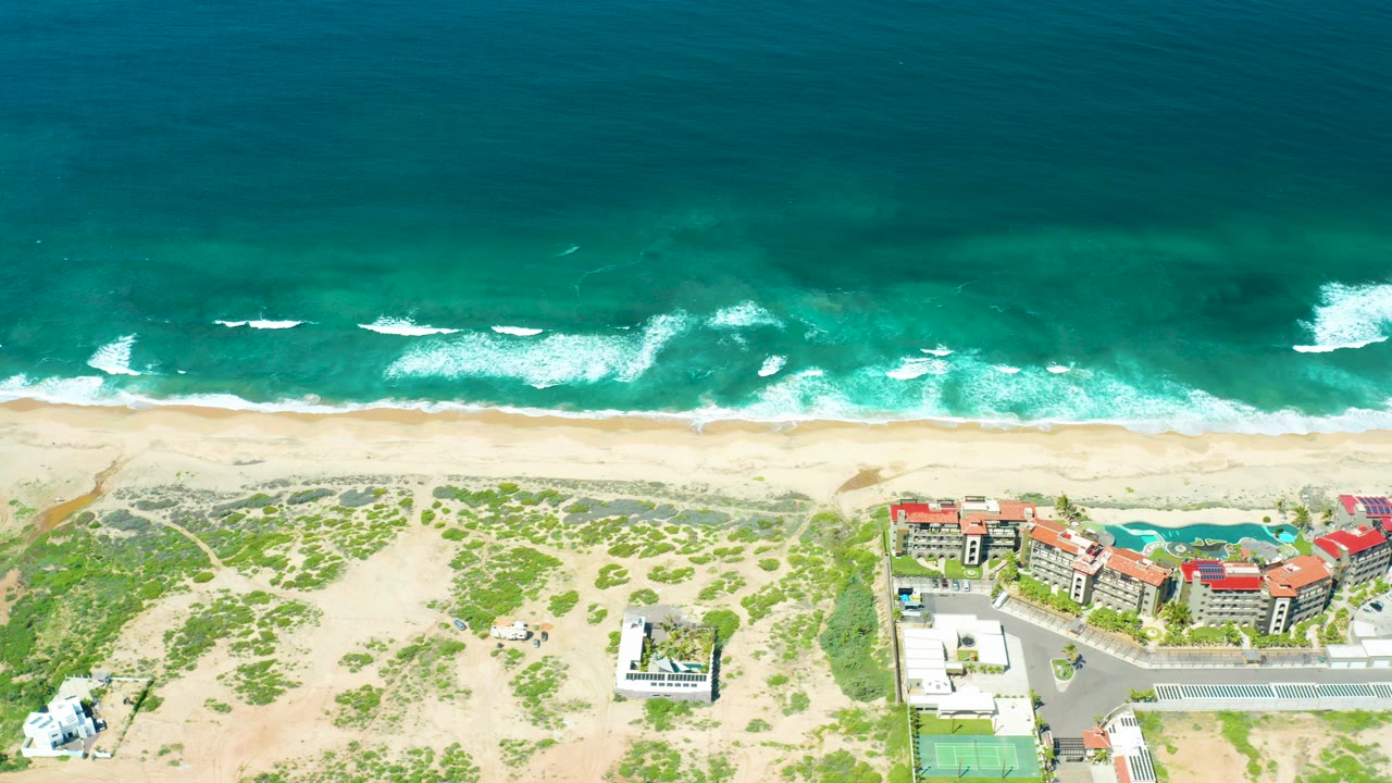 Overhead view of Cerritos Beach in Baja California Sur with turquoise water, white surf lines, pale sand, dune greenery, red-roofed buildings, a road, and no visible people.