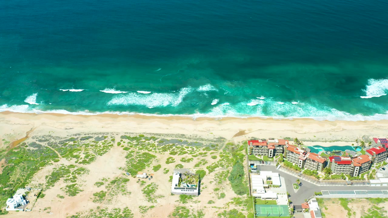 Cerritos Beach aerial frame showing whitecaps closer to shore, a broad sandy strip, scattered dune plants, red roofs, a parking area, and no visible people.