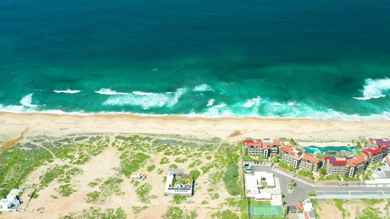 High drone view over Cerritos Beach with teal ocean at the top, foamy wave breaks on the sand, dune vegetation below, red-roofed buildings, paved roads, and no visible people.