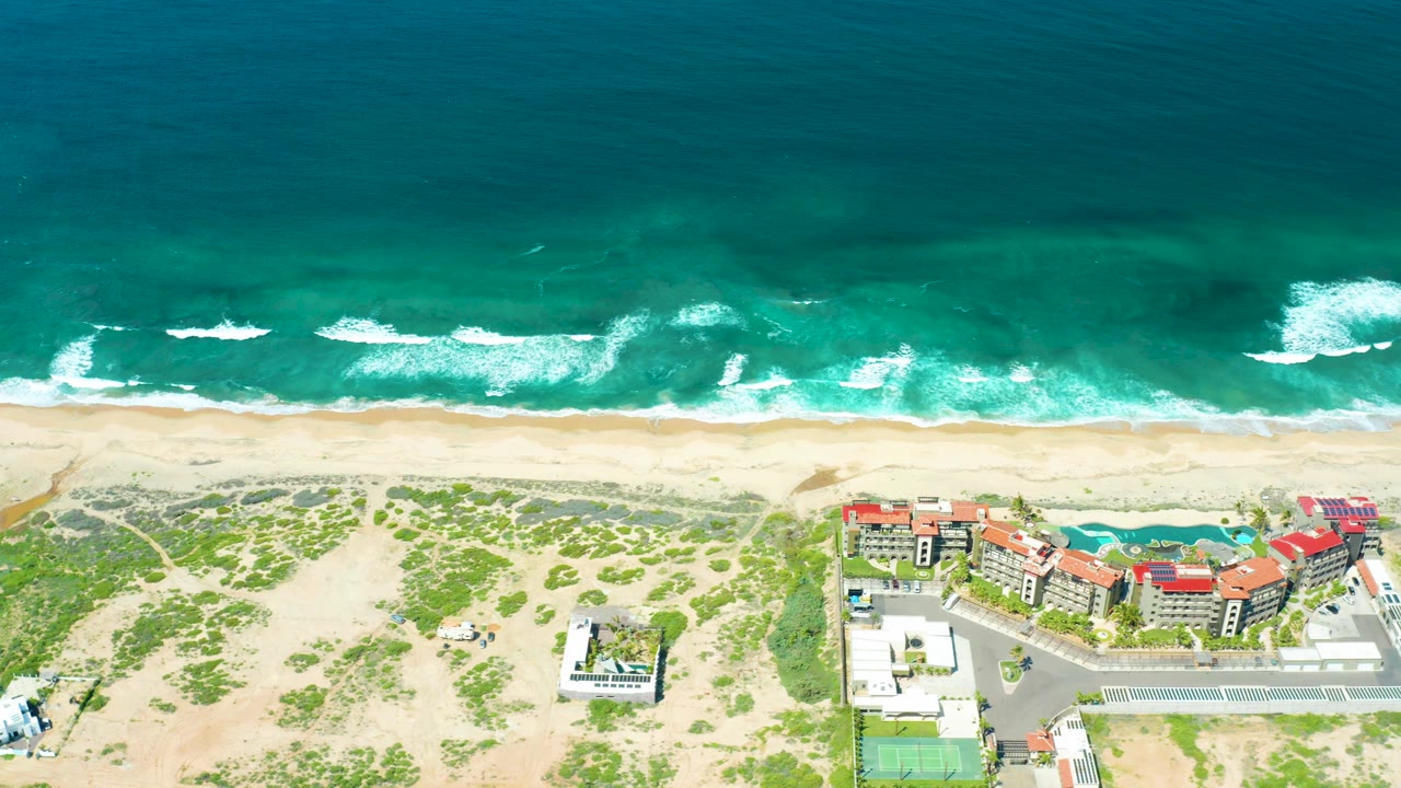 Aerial shoreline view with active whitecaps, pale sand, dunes, and buildings along the lower frame.