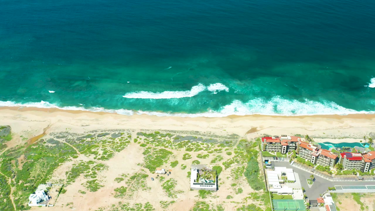 Aerial frame at Cerritos Beach showing white surf advancing toward the sand, scattered green dune growth below, and a cluster of coastal buildings at the lower right, with no visible people.