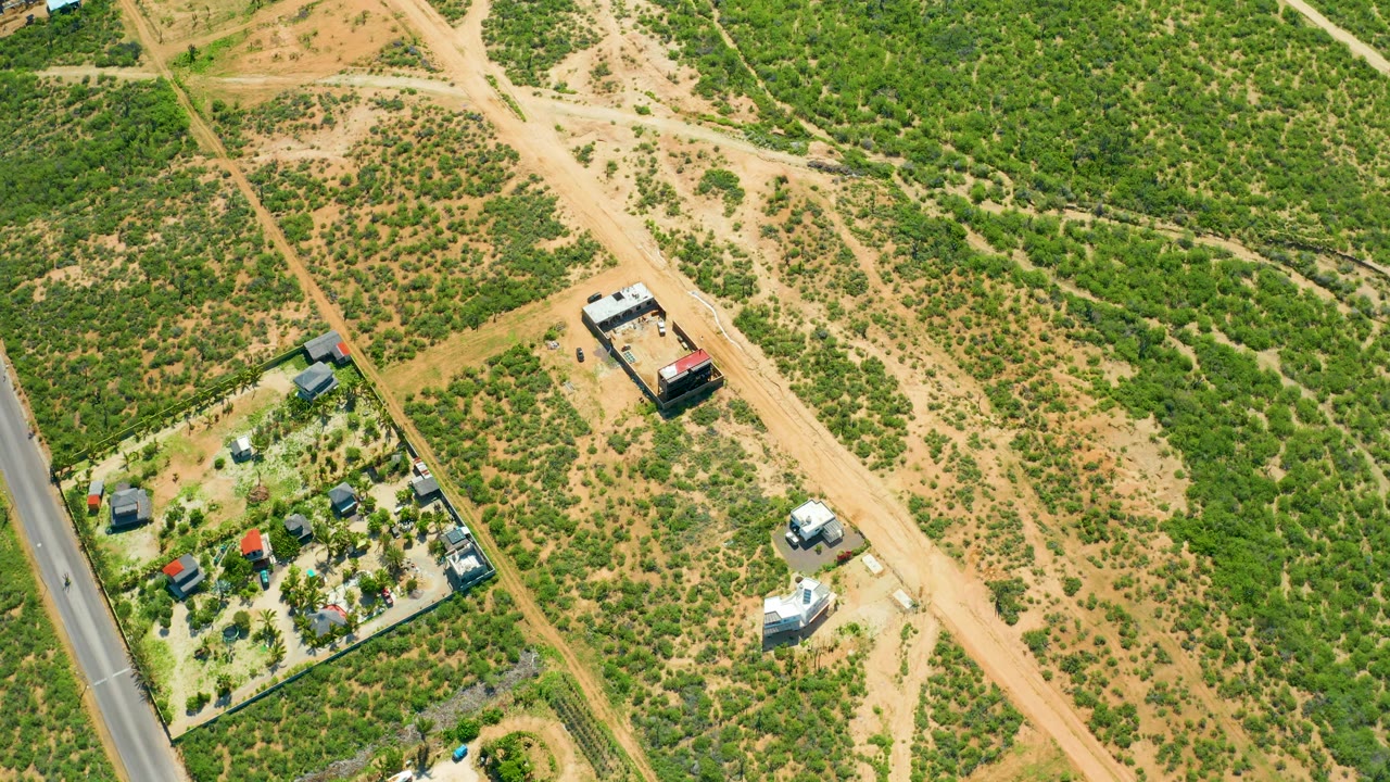 High drone view over Cerritos Beach, Baja California Sur, showing tan dirt roads through patchy green scrub, a paved road at left, scattered buildings, and no visible people.