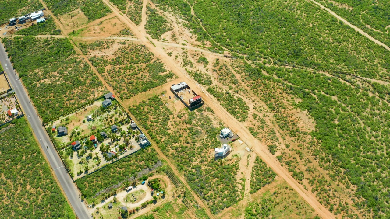 Aerial view in Cerritos Beach, Baja California Sur, with the paved road along the left edge, a diagonal dirt route through green and sandy ground, small structures, vehicles, and no visible people.
