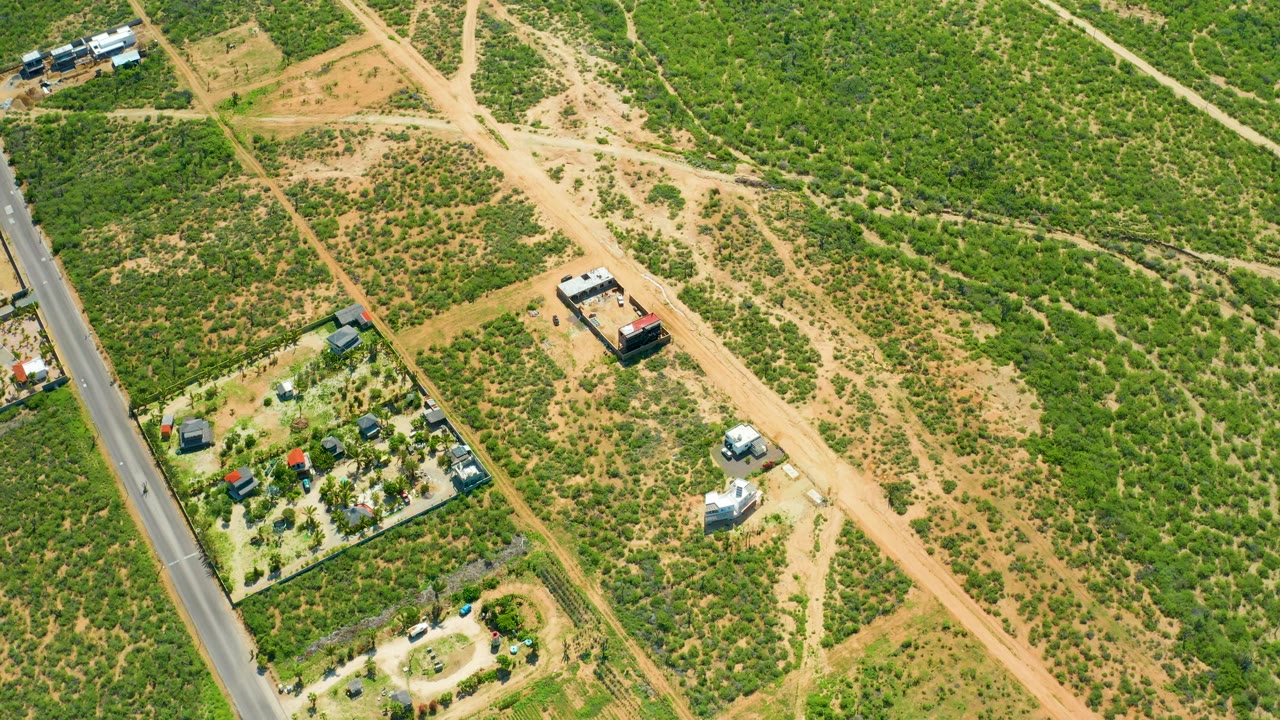Oblique overhead frame from Cerritos Beach, Baja California Sur, showing a pale dirt track cutting across scrubby green terrain, small buildings near the roadside, vehicles on the open road area, and no visible people.