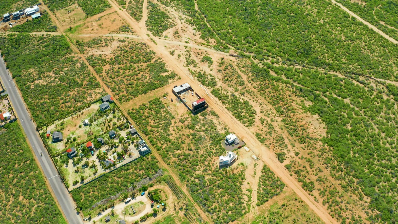 Aerial frame of a rural dirt road crossing green scrub with buildings and vehicles.
