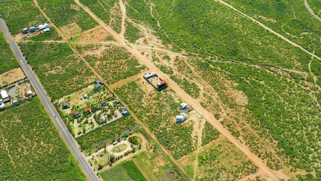 Aerial frame over Cerritos Beach, Baja California Sur, with green scrubland, tan dirt paths, a paved road at left, small properties, and no visible people.