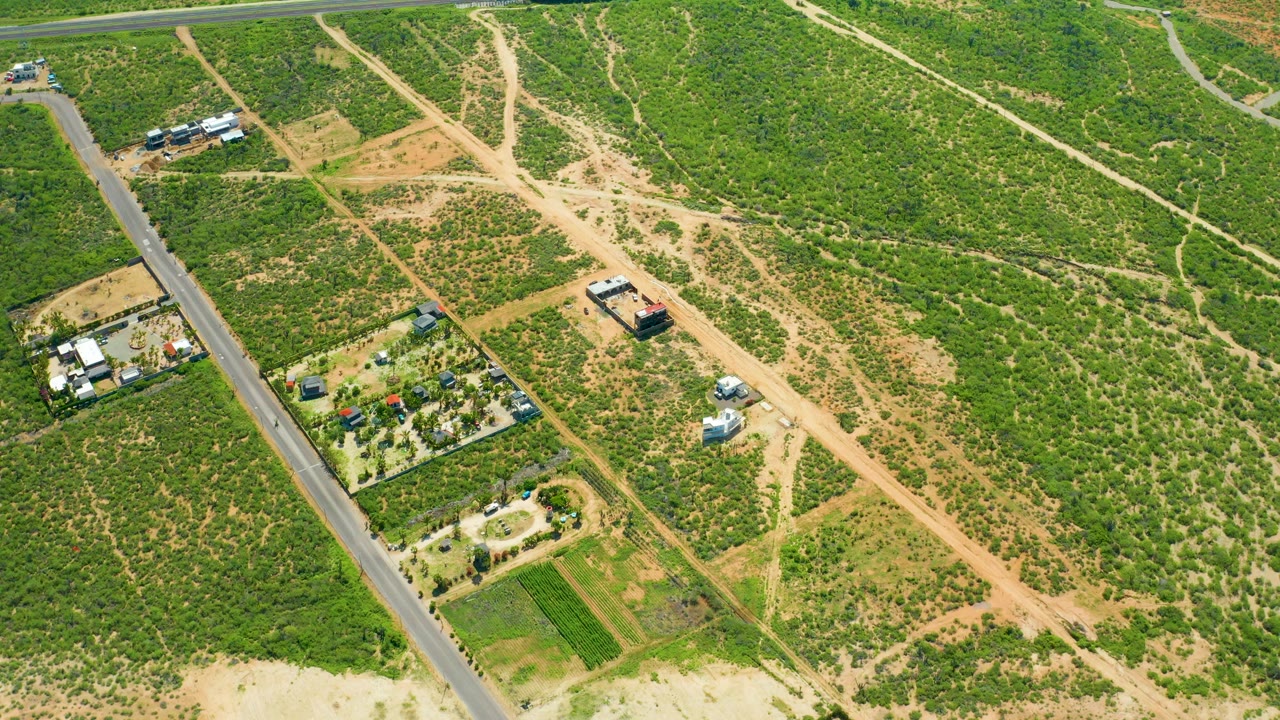 Aerial frame over Cerritos Beach, Baja California Sur showing intersecting tan dirt roads, green scrubland, a paved road along the left side, small structures, cultivated plots, and no visible people.