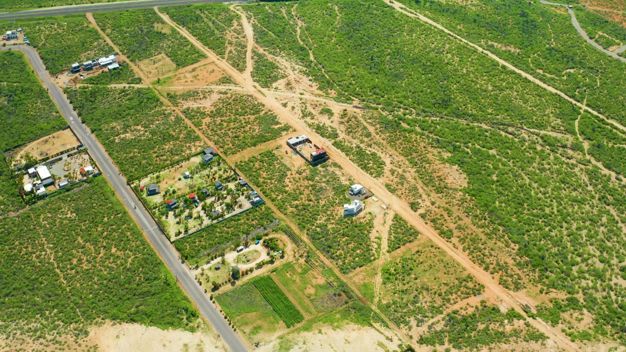Aerial frame over Cerritos Beach, Baja California Sur with the dirt-road junction slightly shifted in view, vehicles visible as small shapes on the track, green fields, rural lots, and no visible people.