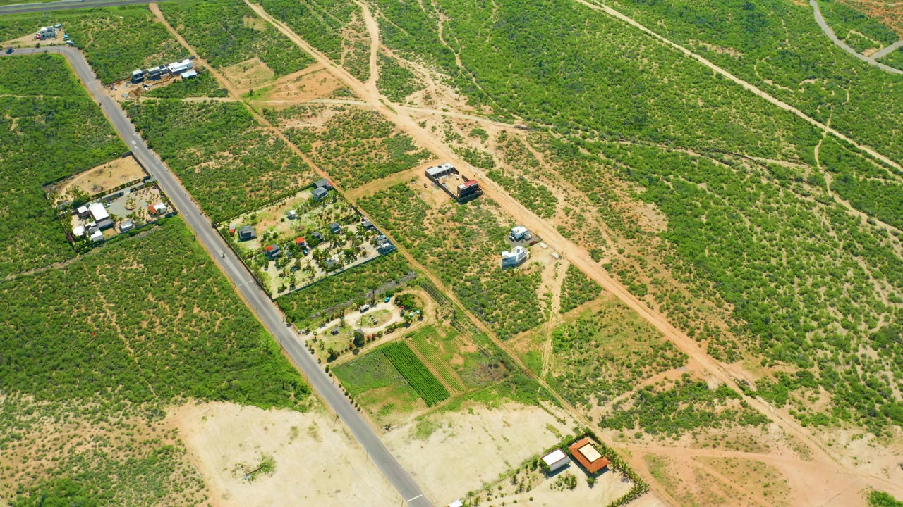 Aerial frame of a car on a country road beside green fields, dry dirt paths, and scattered small structures.