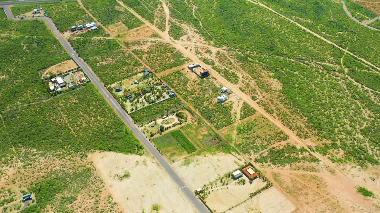 Aerial frame over Cerritos Beach, Baja California Sur showing a paved road on the left, intersecting dirt paths, green scrubland, sandy lots, scattered houses, small vehicles, and no visible people.