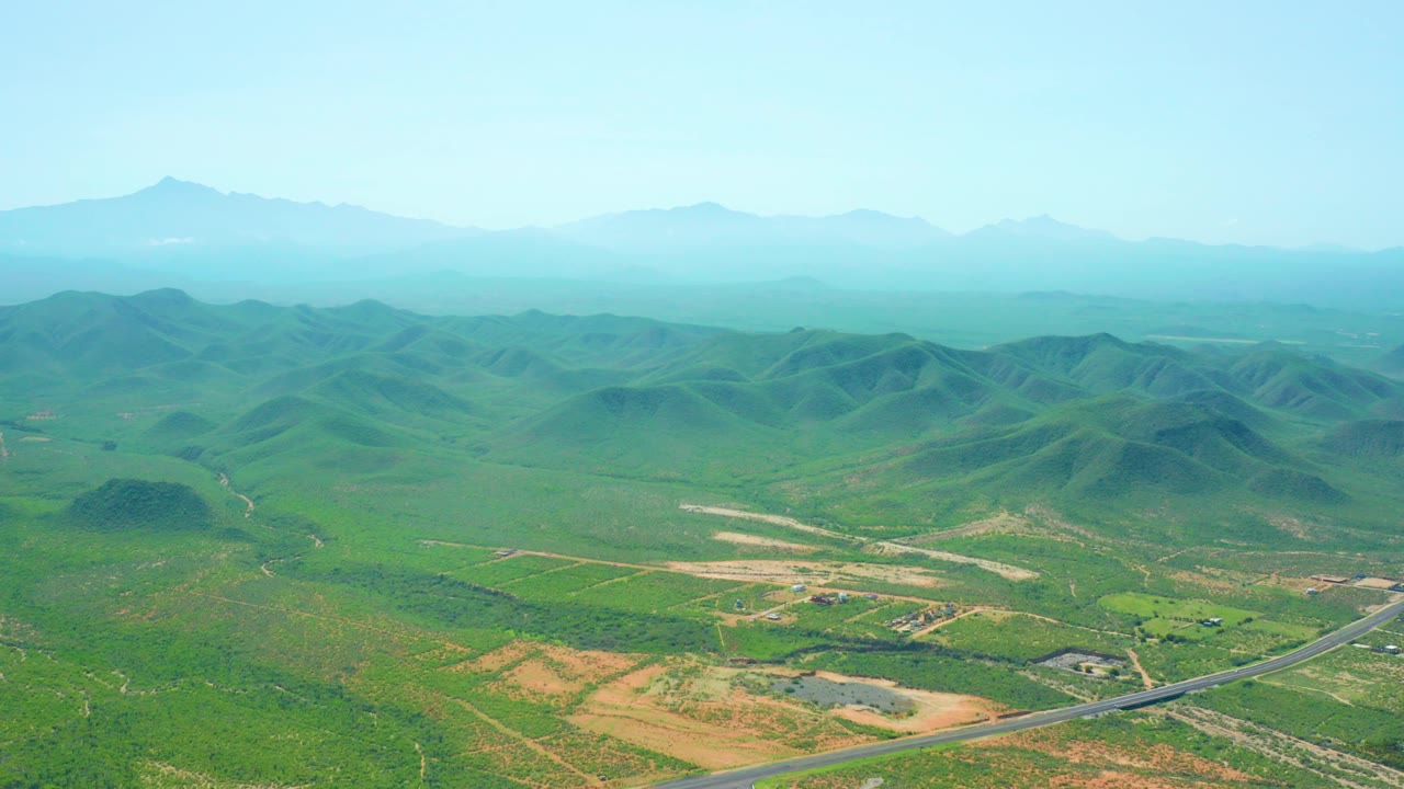 Aerial view over Cerritos Beach, Baja California Sur countryside with green hills, tan fields, a road at the bottom edge, distant mountains, clear sky, and no visible people.