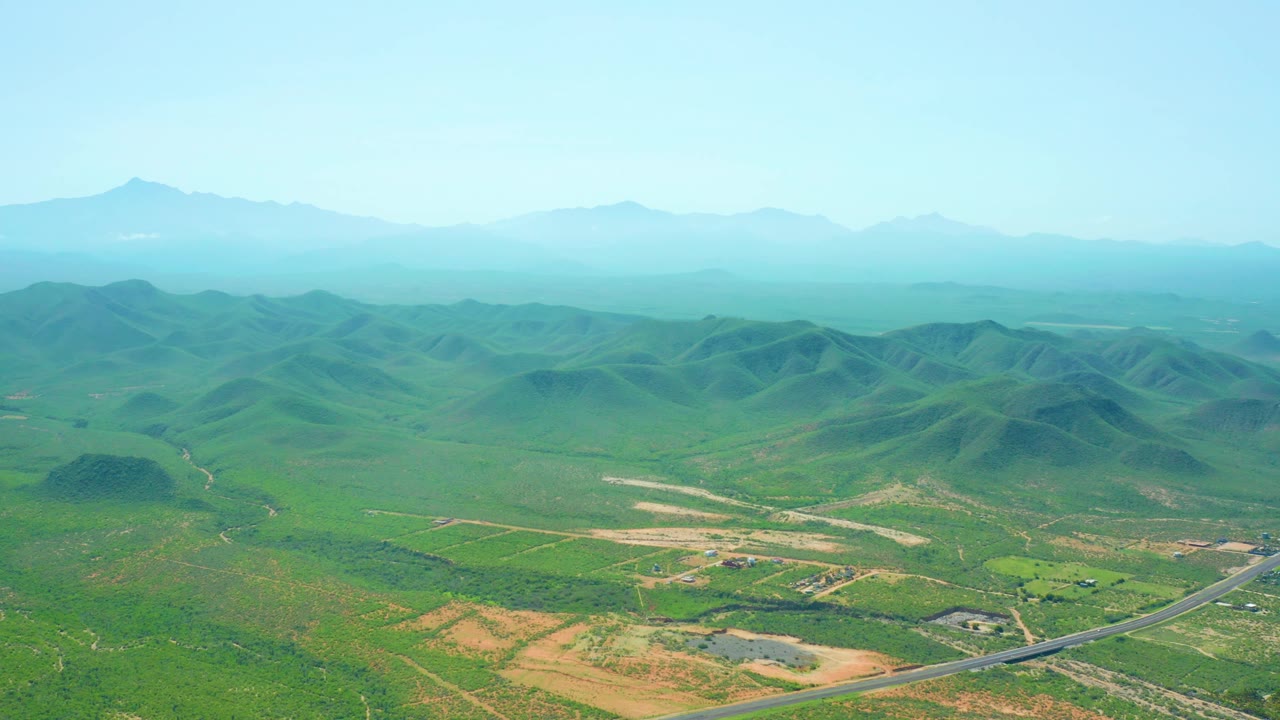 Aerial view of Cerritos Beach, Baja California Sur countryside showing a road crossing the lower right, patchy tan fields, green hill clusters, distant mountain ridges, and no visible people.