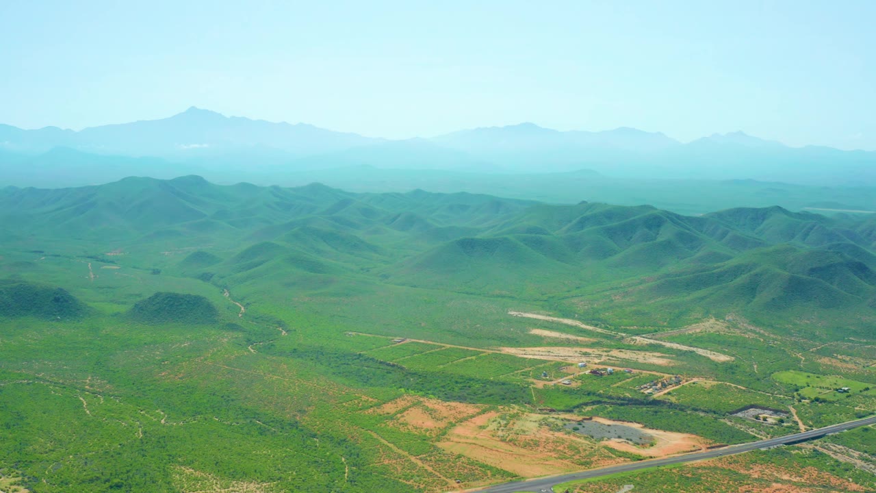 Early aerial view of Cerritos Beach, Baja California Sur rural terrain, with the winding road low in frame, fields and tracks ahead, hazy mountains behind, and no visible people.